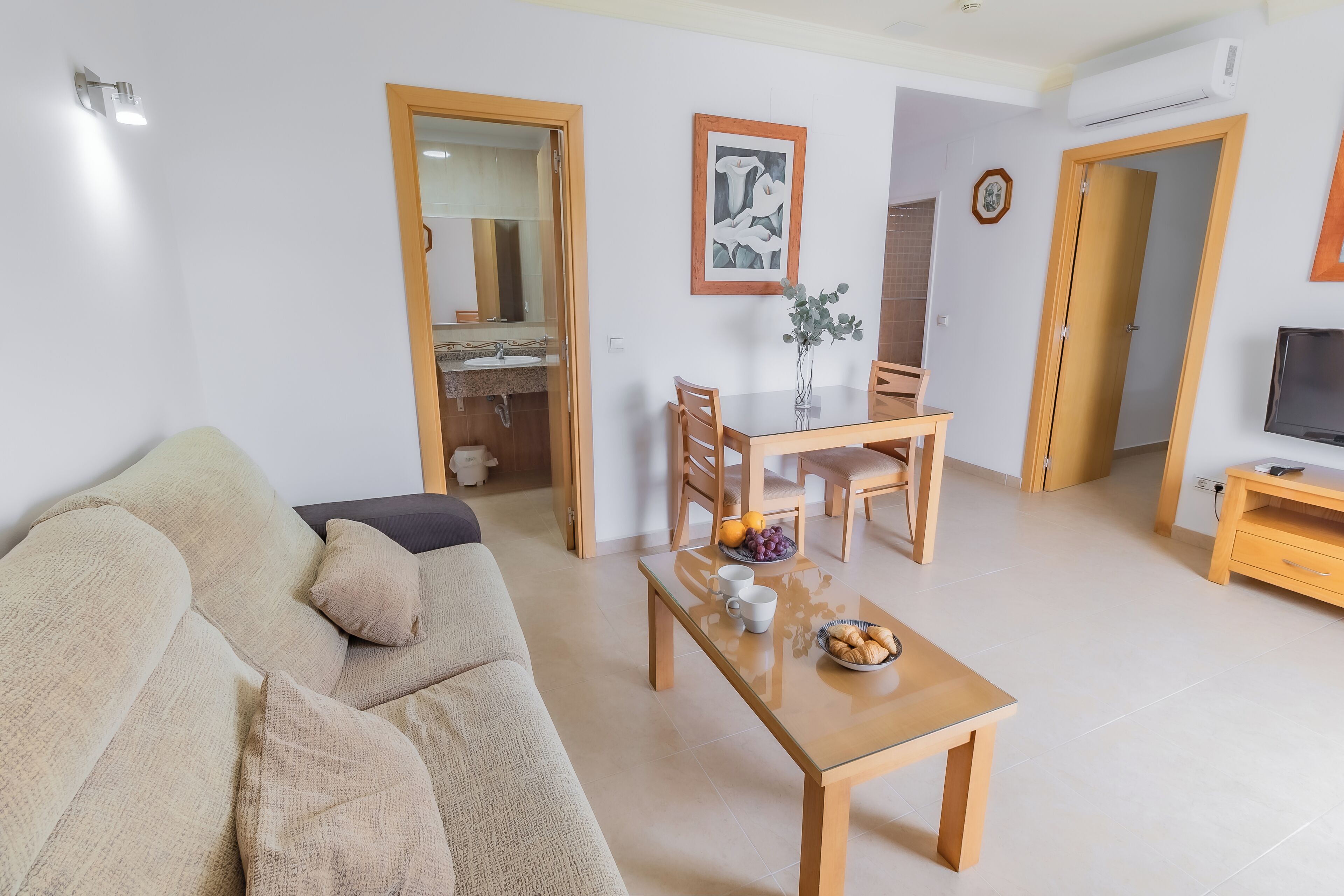Una sala de estar con un sofá, una mesa de centro con frutas y tazas, una zona de comedor con sillas y una mesa, y un mueble para televisión.