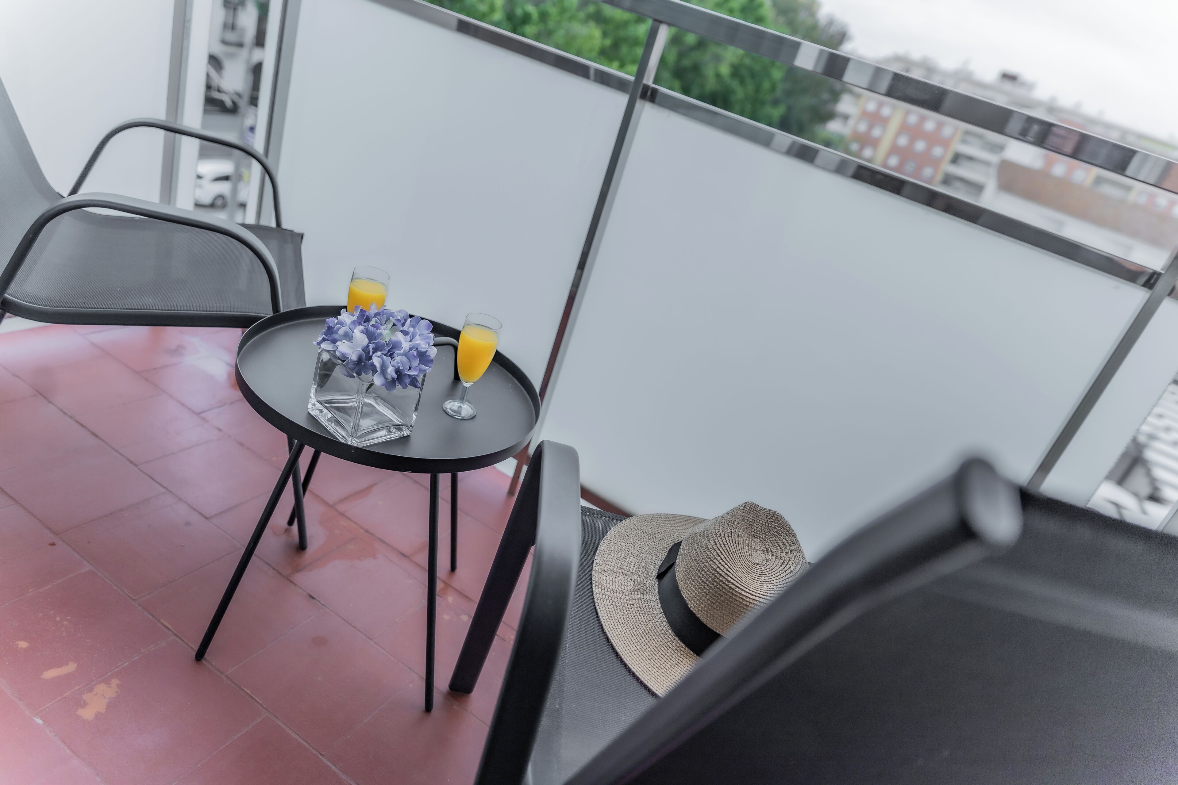 A balcony with a table set for two, featuring a glass of orange juice, a glass of water, and a vase with flowers.