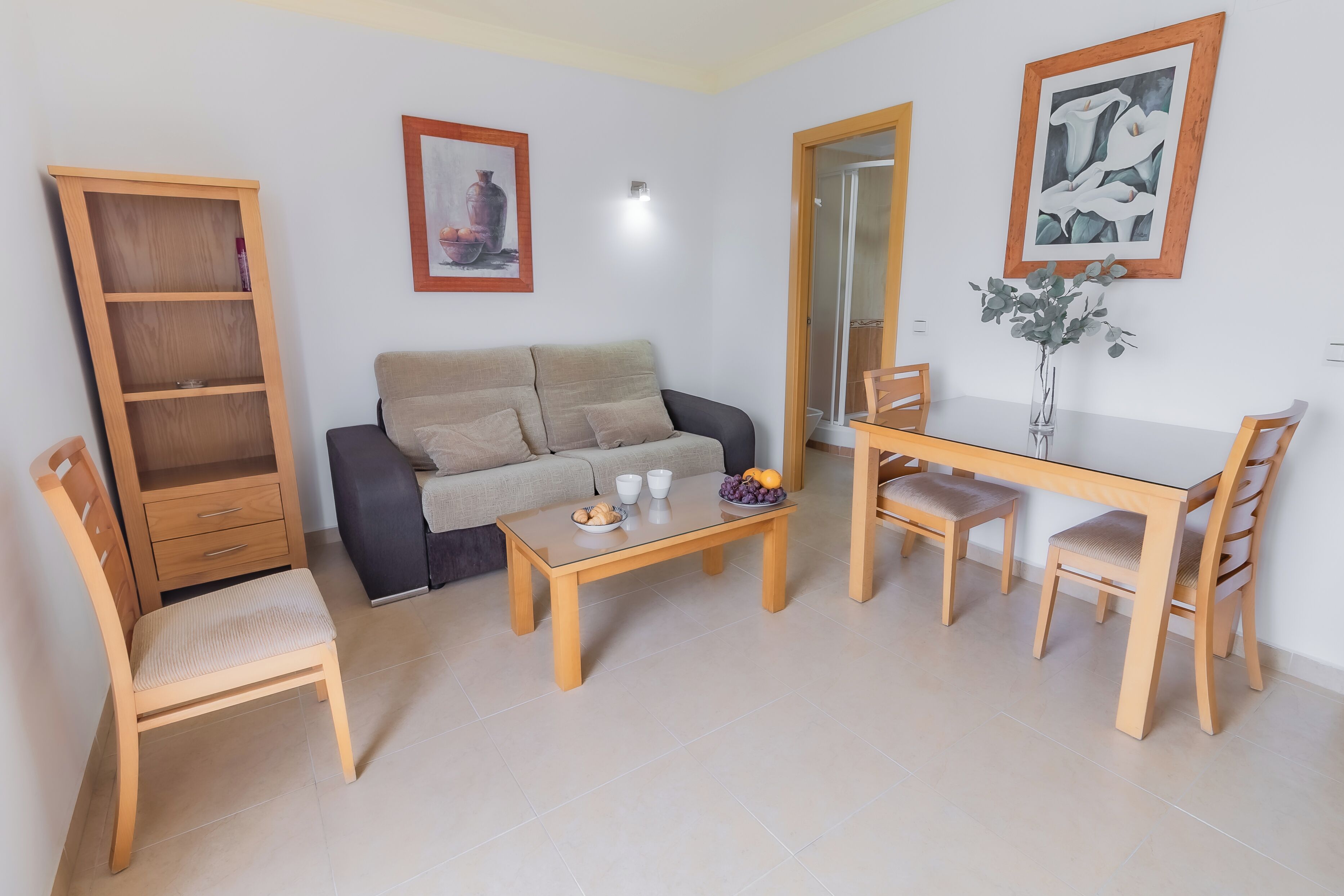 A living room with a sofa, a wooden table, a dining table with chairs, a bookshelf, and two framed pictures on the wall.