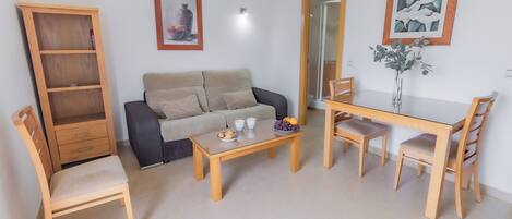 A living room with a sofa, a wooden table, a dining table with chairs, a bookshelf, and two framed pictures on the wall.