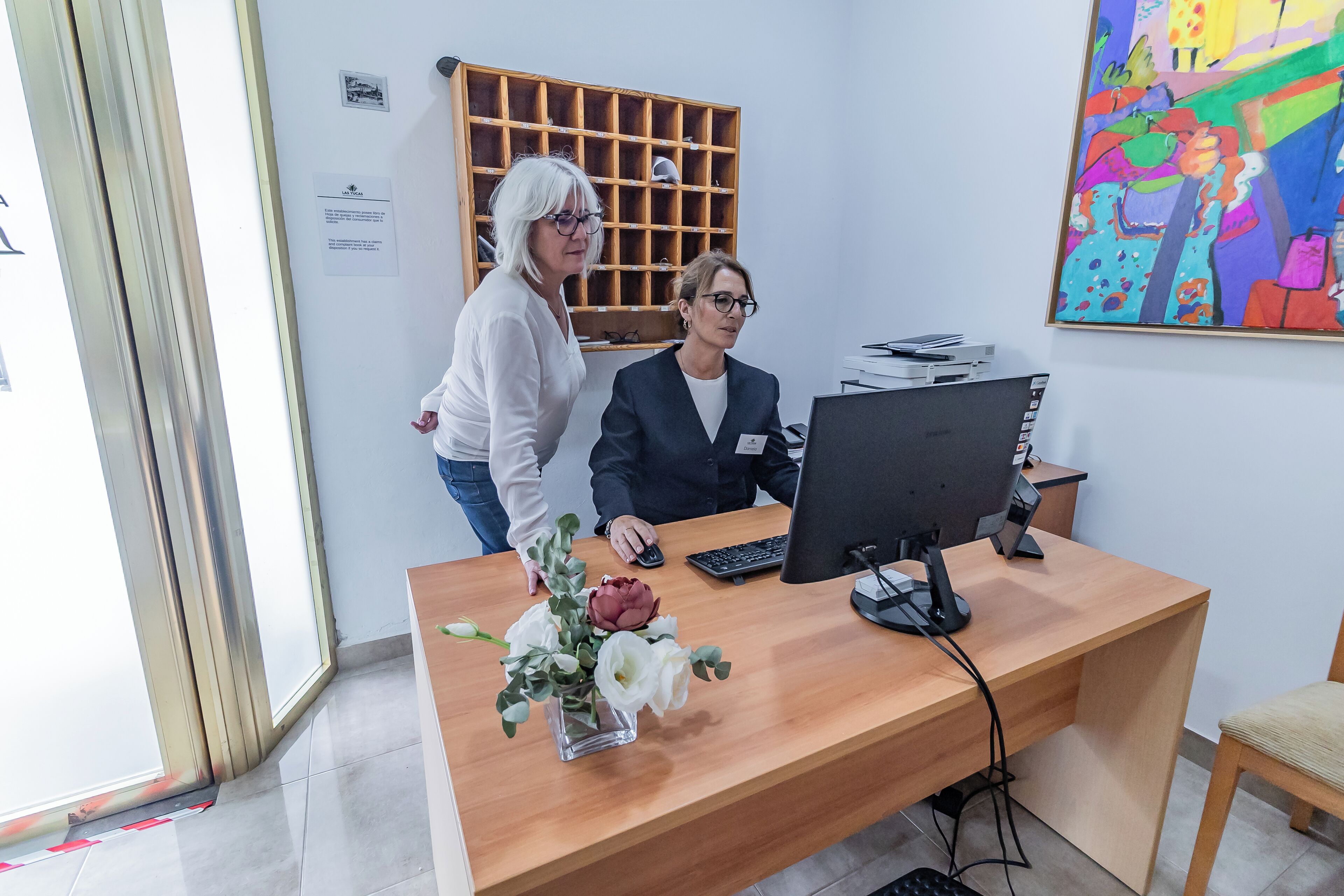 En receptionsdisk med en dator, en vas med blommor och en färgstark tavla på väggen.