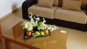 Una sala de estar con un sofá, una mesa baja con fruta y flores, y una zona de cocina con armarios y un fregadero.