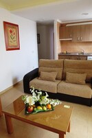 Una sala de estar con un sofá, una mesa baja con fruta y flores, y una zona de cocina con armarios y un fregadero.