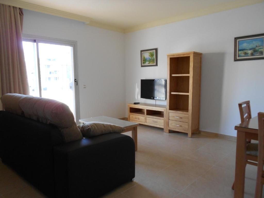 A living room with a sofa, a TV on a wooden stand, a wooden cabinet, and a dining table with chairs.