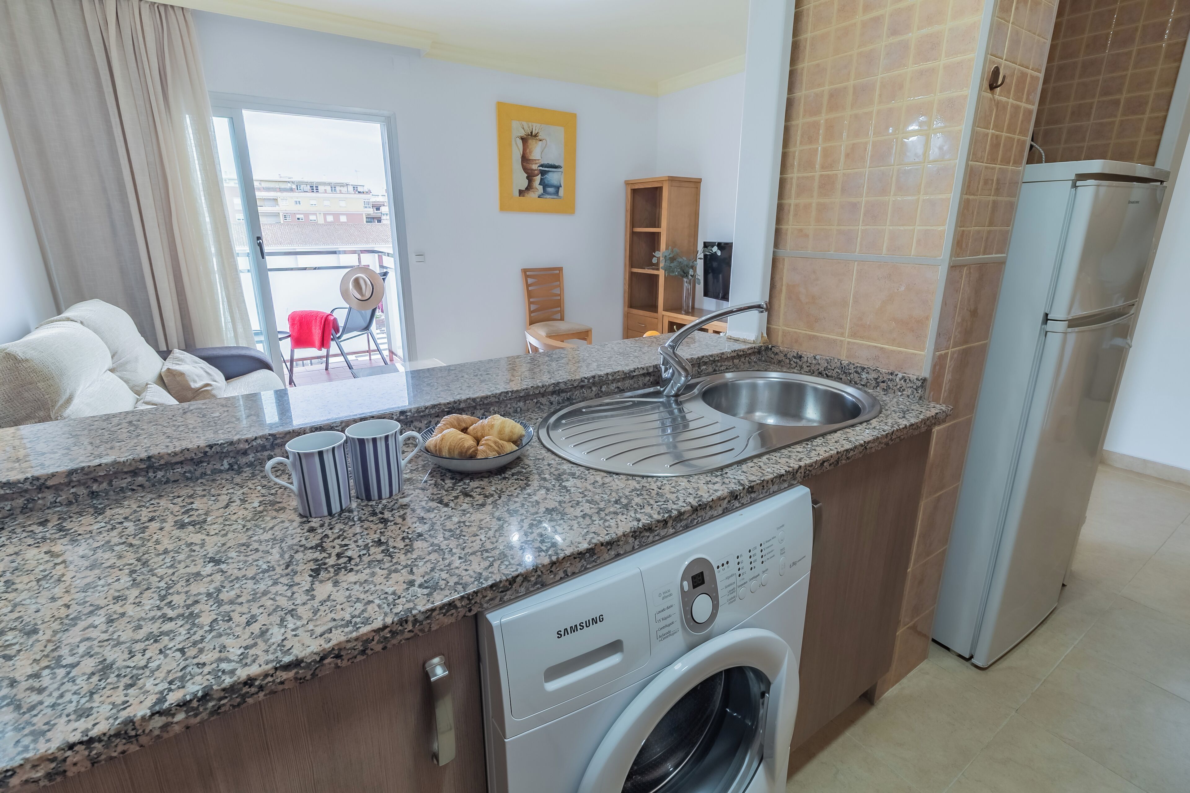 A kitchen with granite countertops, a stainless steel sink, a Samsung washing machine, and a refrigerator.