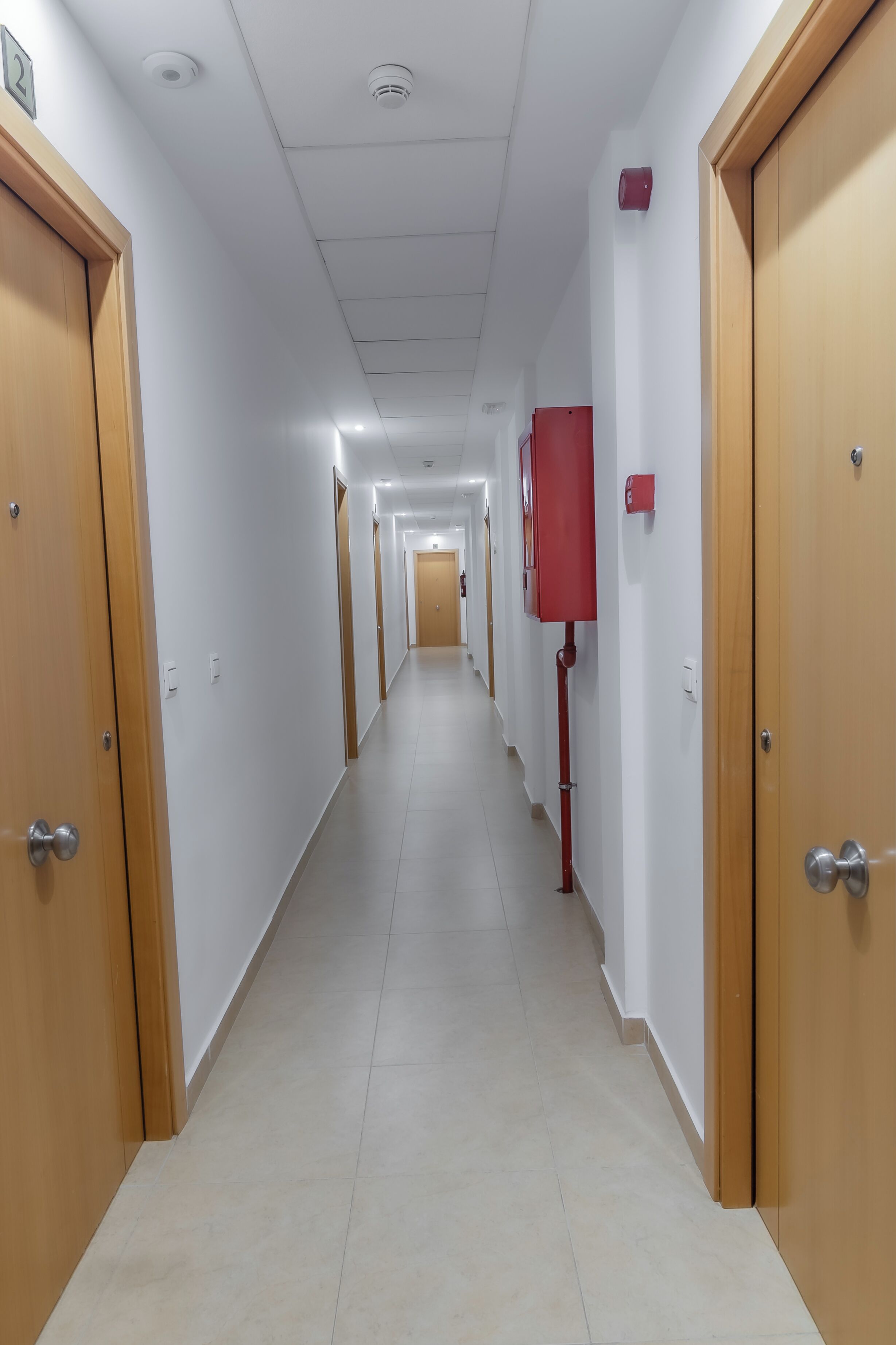 Un pasillo largo y estrecho con puertas de madera a ambos lados, paredes blancas y suelo de baldosas.