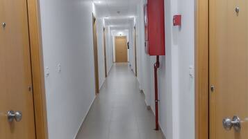 Un pasillo largo y estrecho con puertas de madera a ambos lados, paredes blancas y suelo de baldosas.