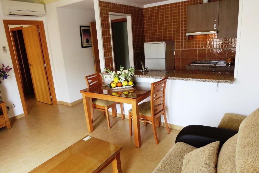 A dining area with a wooden table and chairs, a fruit bowl, and a kitchen in the background.