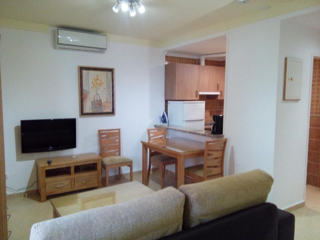 Una sala de estar con televisor, muebles de madera y una cocina al fondo.