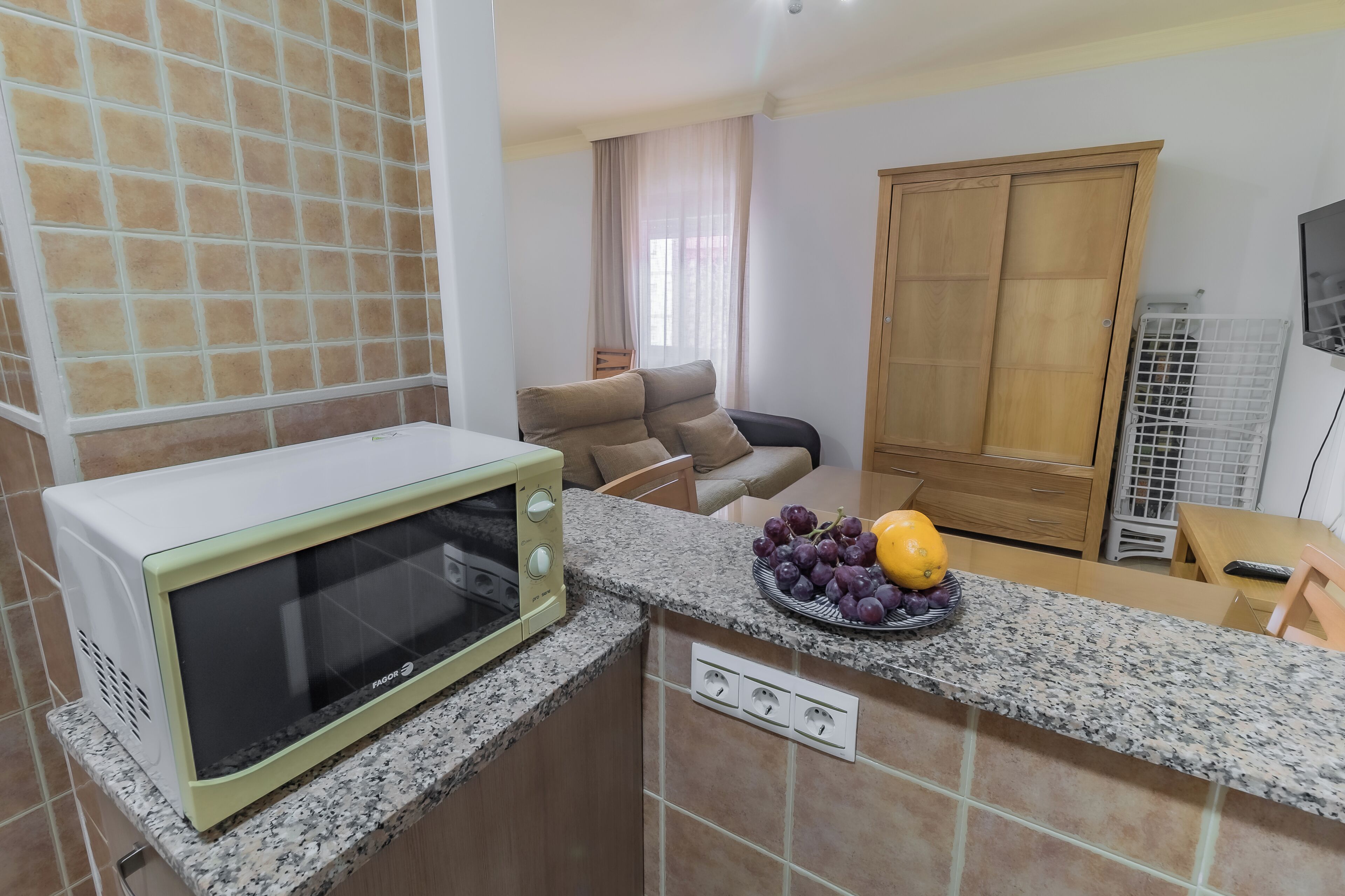 A kitchen with a microwave, granite countertop, and a fruit bowl with grapes and a melon.