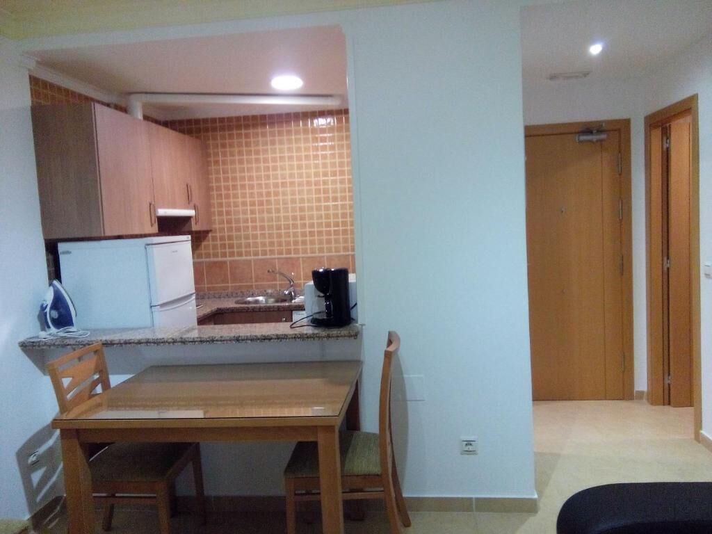 A small kitchen with a refrigerator, stove, and coffee maker. There is a wooden table and chairs.