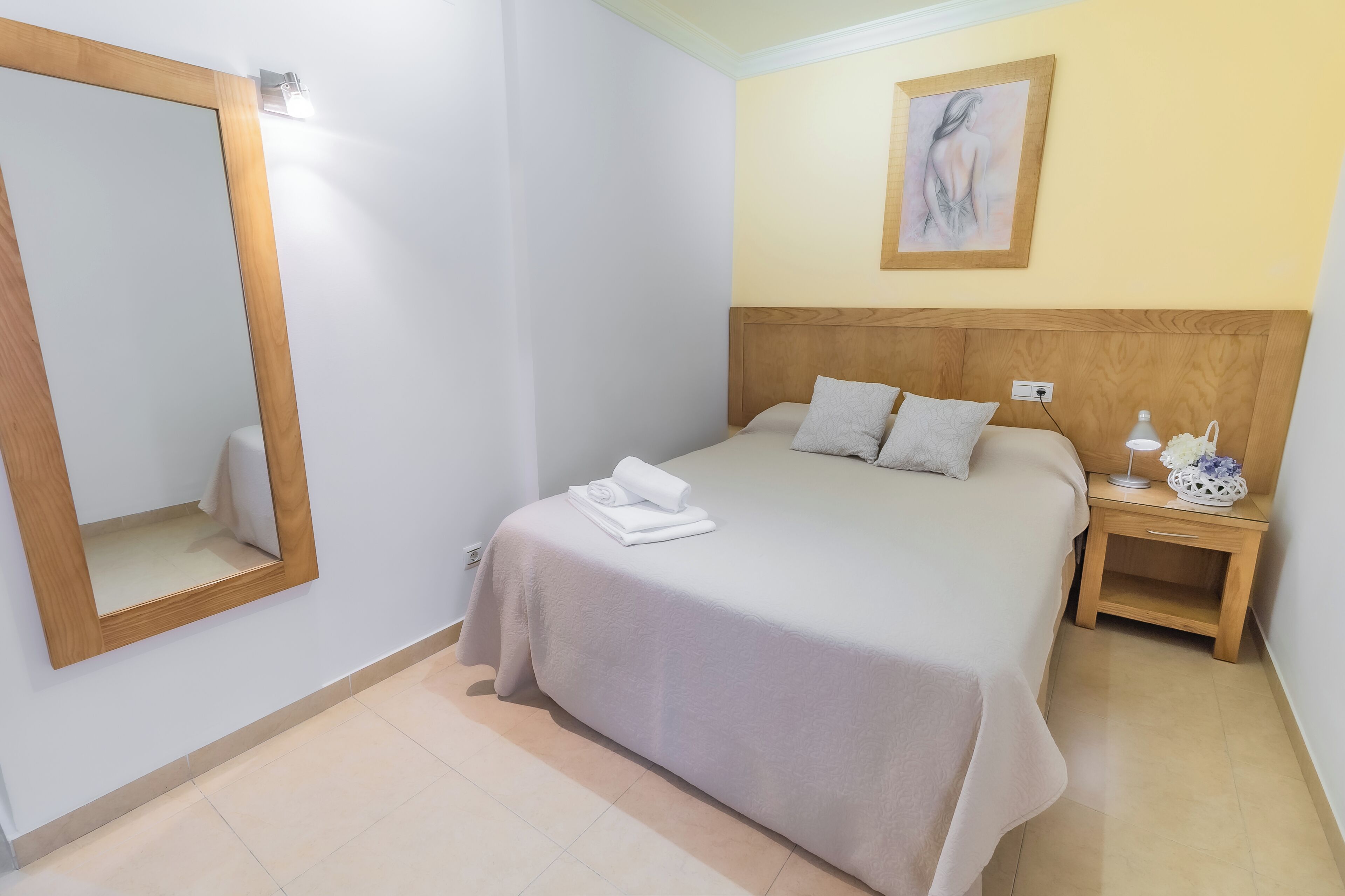 A neatly made bed with a light-colored bedspread and pillows, a wooden nightstand with a lamp and decorative items, a large mirror on the wall, and a painting above the bed.