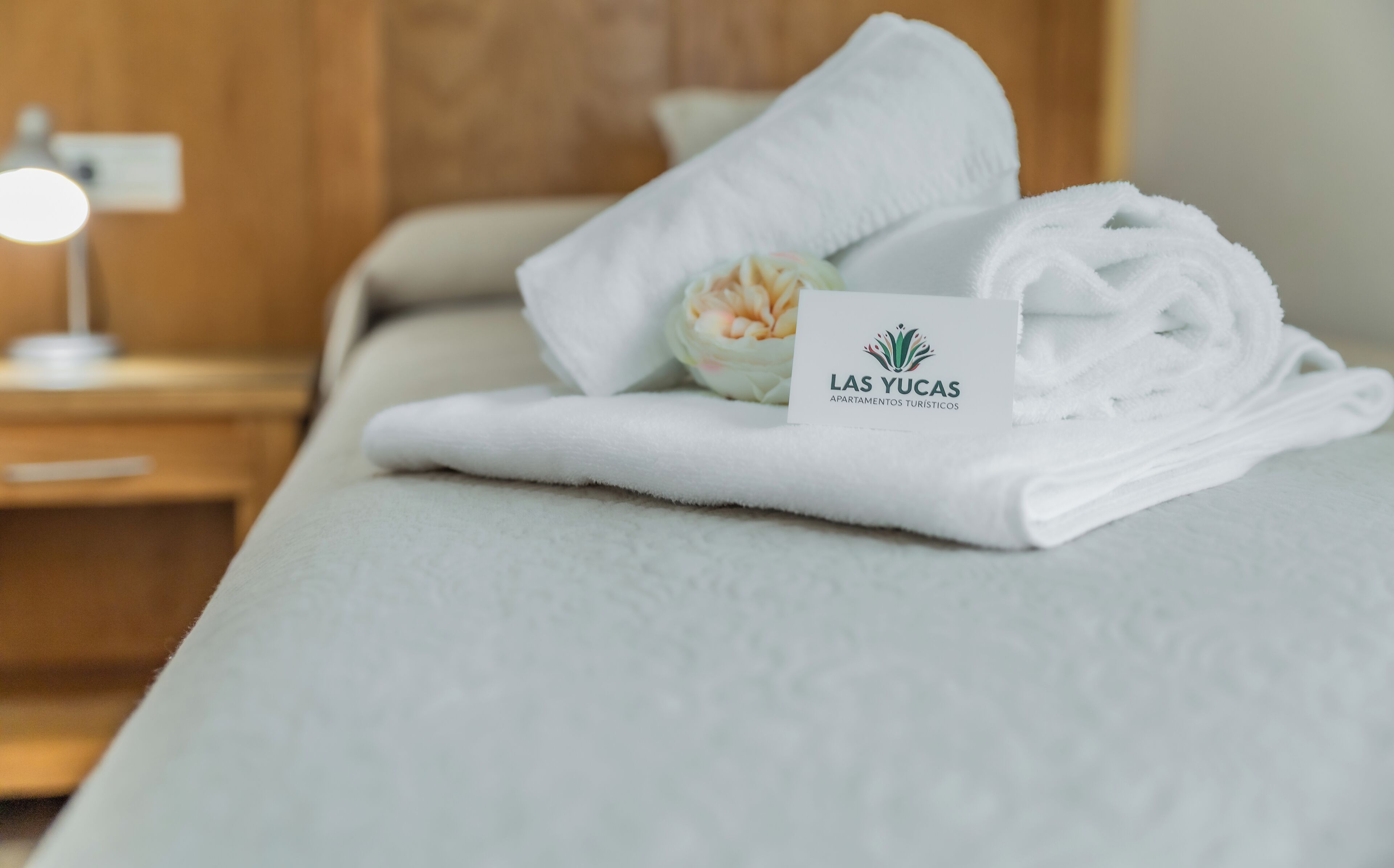 A neatly made bed with a folded white towel and a card labeled "Las Yucas Apartamentos Turísticos" on it.