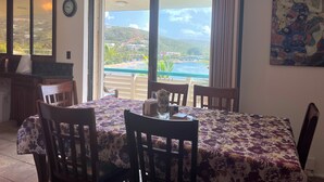 A dining area with a table set for four, a ceiling fan, and a view of a coastal landscape through large windows. - Our Home is Your Home in St Thomas, ~USVI~ (Charlotte Amalie)