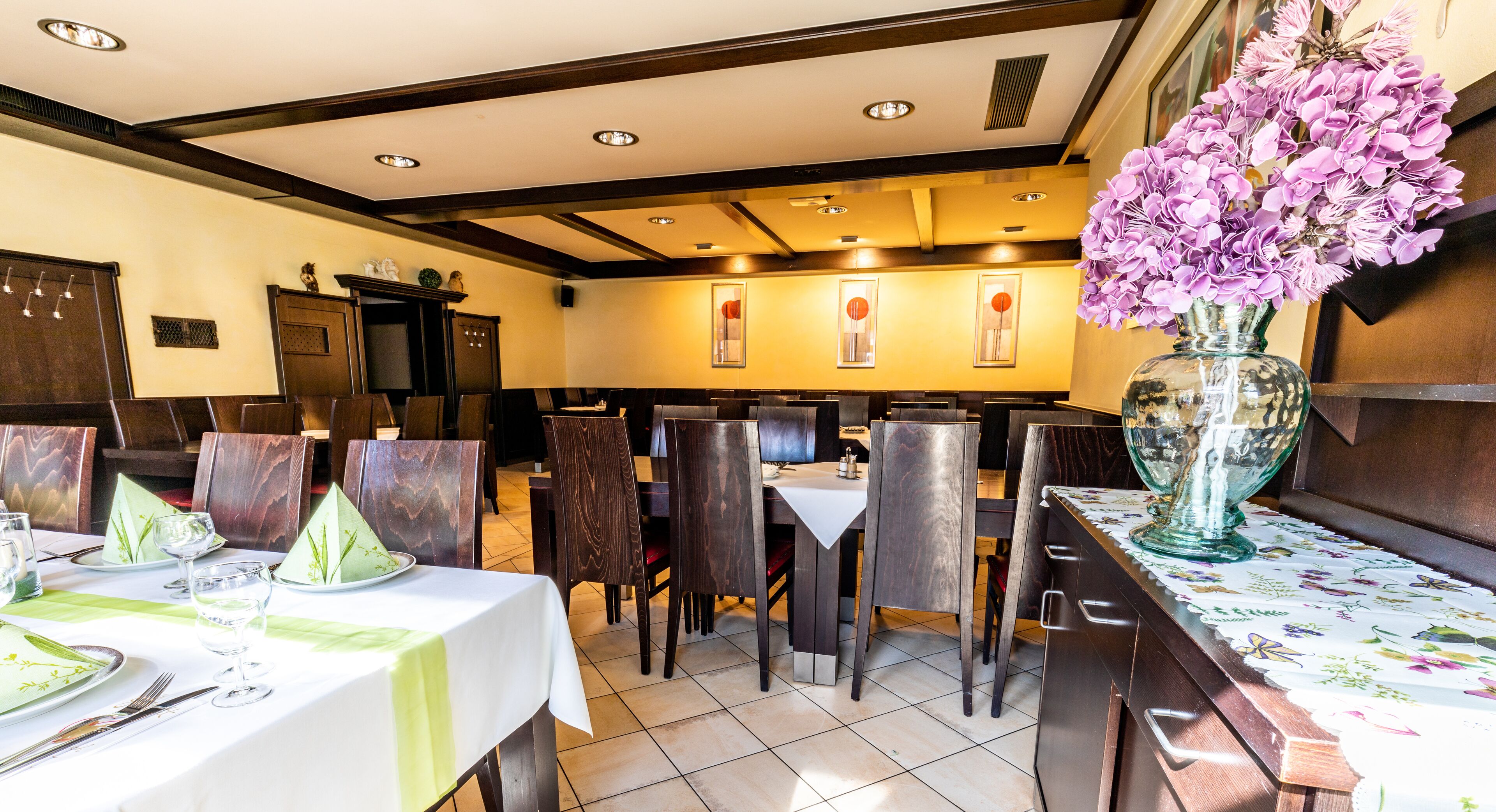 Ein Restaurant mit Holztischen und -stühlen, einer großen Vase mit lila Blumen und einem Fliesenboden.