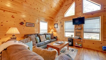 A cozy cabin interior with a wooden ceiling, a leather sofa, a television, and a window with a view.