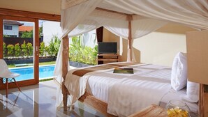 A bedroom with a four-poster bed, a television on a wooden stand, a glass table with a bowl of fruit, and a view of a swimming pool and greenery outside. - Prime location just walking distance to the Boutique shop, Restaurant , Bar (Seminyak)