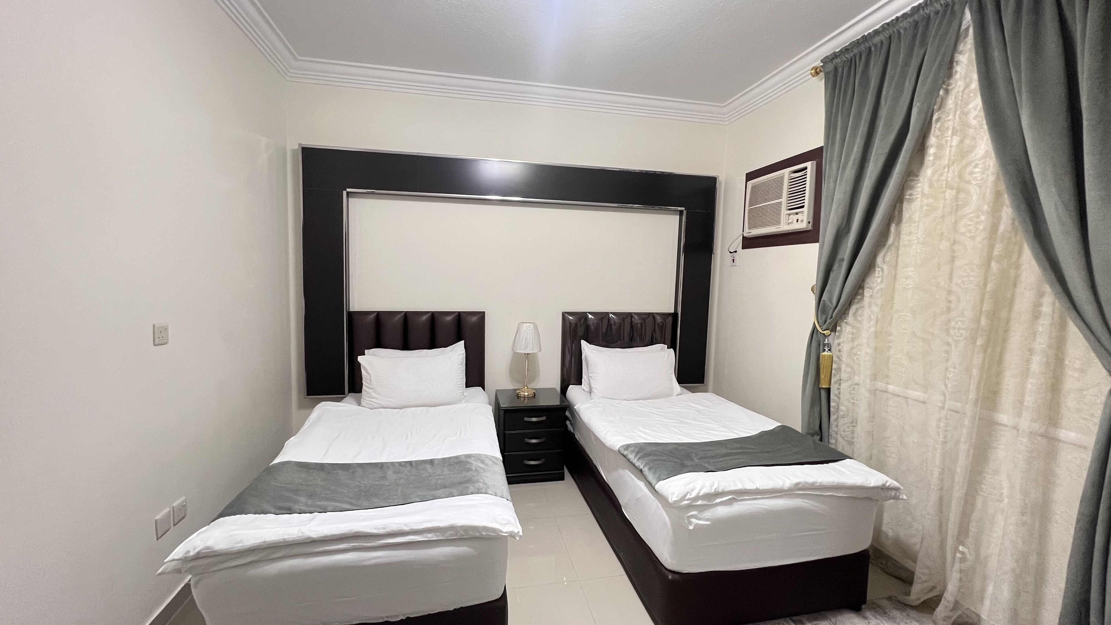 Habitación de hotel con dos camas individuales, una mesita de noche con lámpara, un espejo grande sobre las camas y una ventana con cortinas.