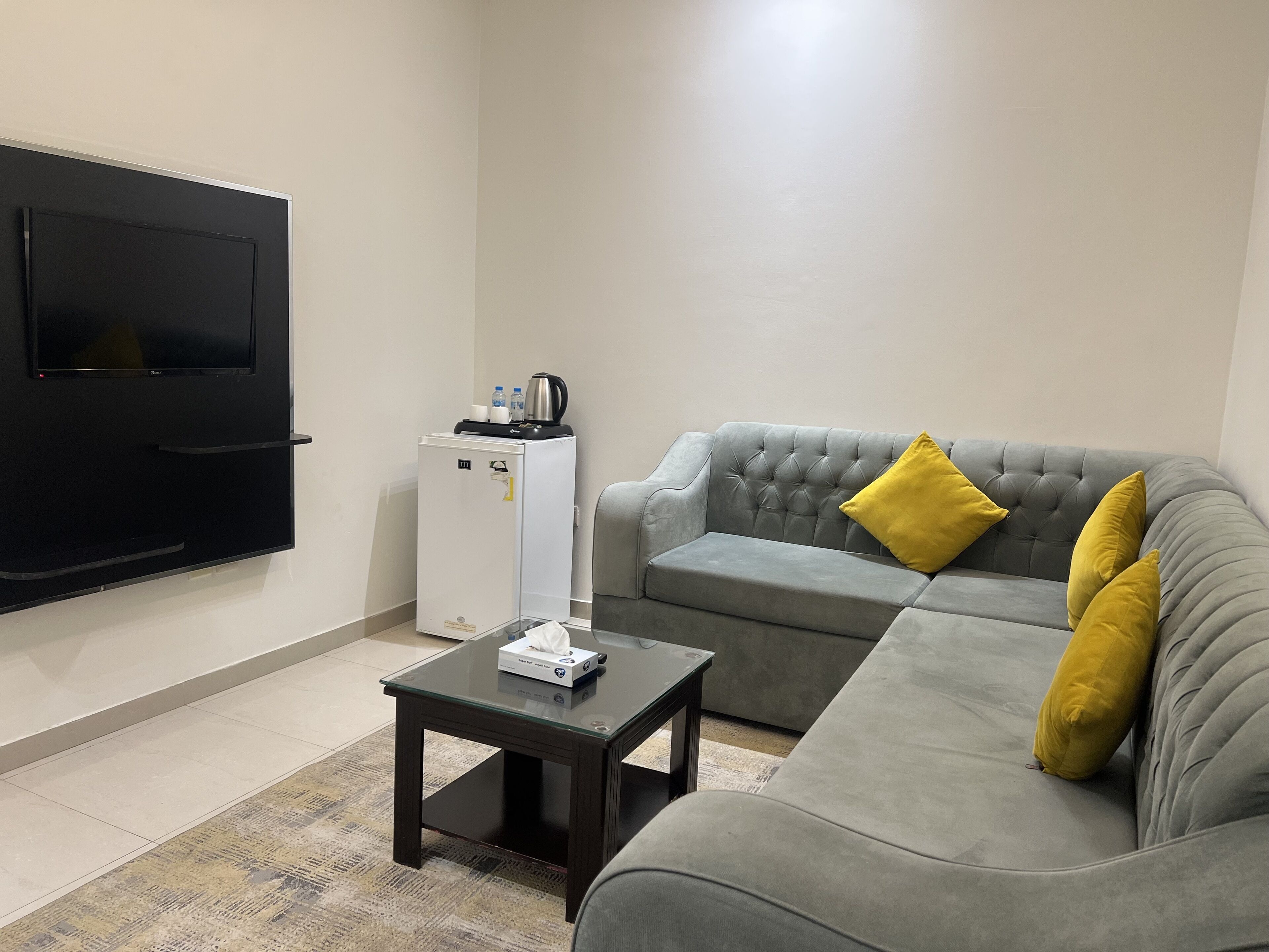 Ein Wohnzimmer mit grauem Sofa, einem kleinen schwarzen Couchtisch, einem weißen Mini-Kühlschrank und einem flachbildfernseher an der Wand.