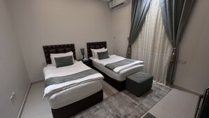 Habitación doble con dos camas, una mesita de noche con lámpara, un pequeño reposapiés y una ventana con cortinas.