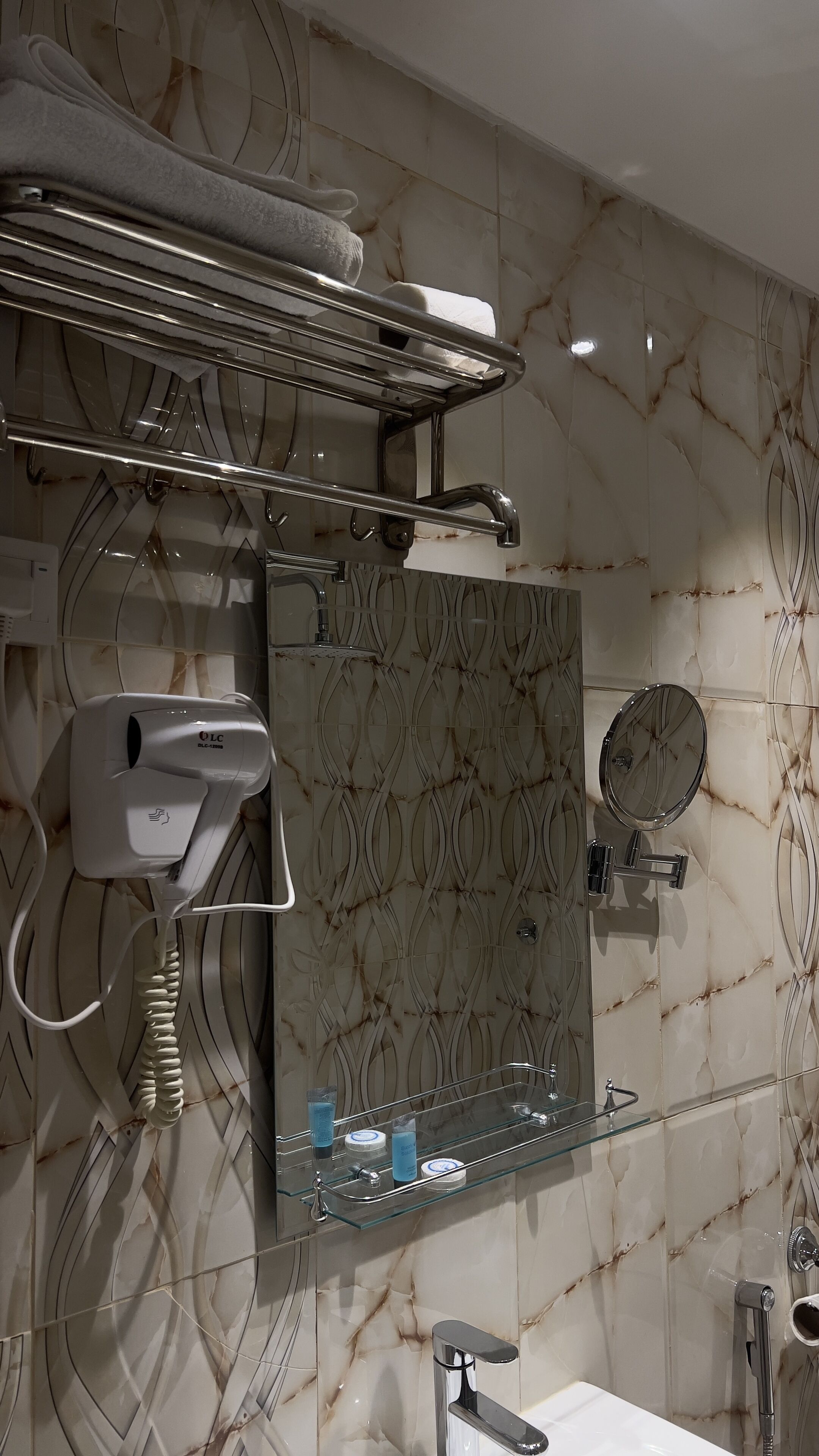 A bathroom with a marble wall, a towel rack, a hairdryer, a mirror, and a glass shelf with toiletries.