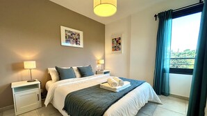 Un lit bien fait, avec des oreillers, une lampe de chevet et une pile de serviettes. Deux lampes murales et un tableau encadré sont accrochés au mur, au-dessus du lit.