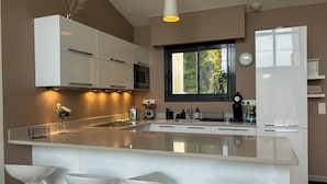 Une cuisine moderne dotée d’un îlot central, de meubles blancs et d’une fenêtre donnant sur un espace verdoyant, ainsi qu’une horloge murale.