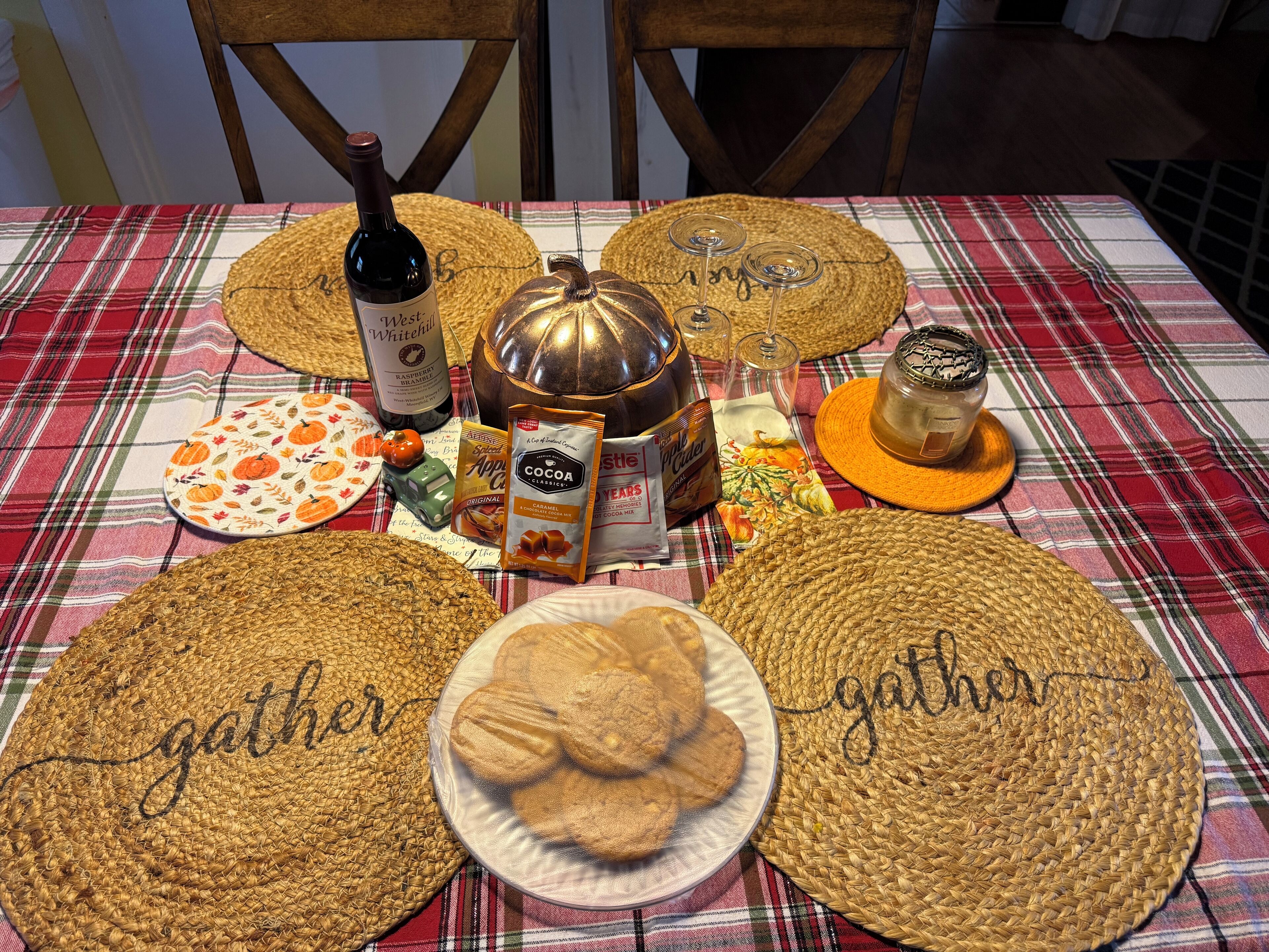 A table set with various food items, including a bottle of wine, a pumpkin, and a plate of cookies, on a plaid tablecloth.
