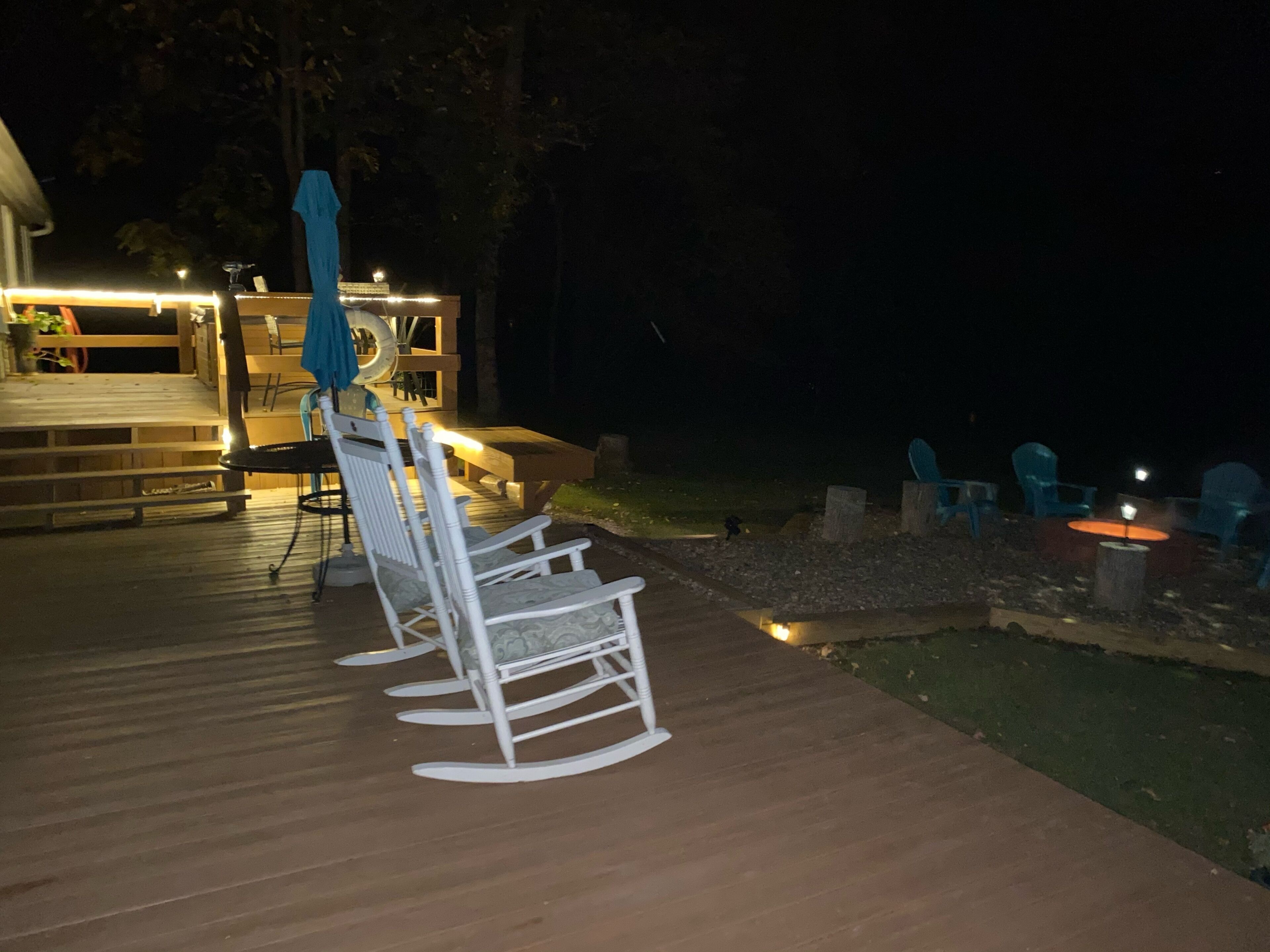 Une terrasse dotée de chaises berçantes blanches et d’une table, entourée d’une autre terrasse en bois et d’un petit espace aménagé pour un foyer.
