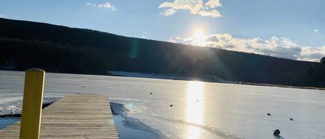 A frozen lake with a wooden dock extending into the water, surrounded by forested hills.