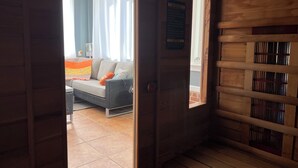Een houten sauna met een bank, een raam met gordijnen, en uitzicht op een zithoek met een bank en een kleurrijk plaid.
