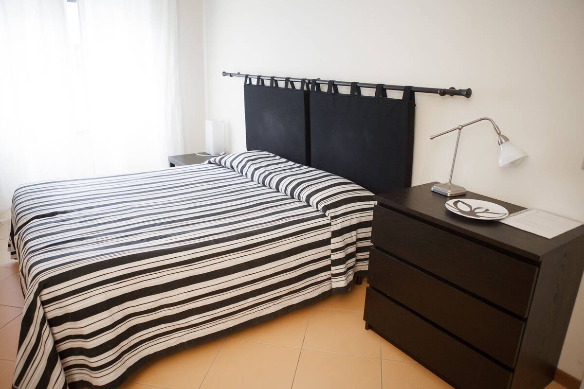 A neatly made bed with striped bedding, a bedside table with a lamp, and a dark dresser.