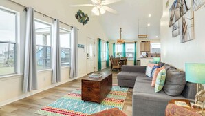 A spacious living room with a gray sofa, a wooden coffee table, and a colorful rug. There is a dining area with a chandelier and teal curtains. The kitchen is visible in the background with wooden cabinets and a hanging light fixture. - Jamaica Beach Gem - Huge Deck, Fenced, Pups Welcome! (Jamaica Beach)