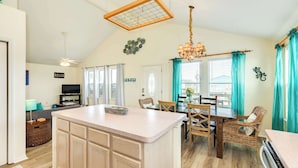 A spacious kitchen with a central island, wooden cabinets, and a dining area with wicker chairs and a table. - Jamaica Beach Gem - Huge Deck, Fenced, Pups Welcome! (Jamaica Beach)