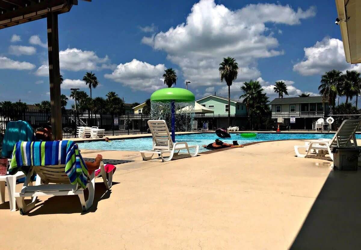 Un espace aménagé au bord de la piscine, avec des chaises longues, un élément décoratif aquatique et une vue sur une maison et des palmiers.