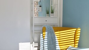 Un sillón amarillo con un plaid suave, una mesita redonda con fruta y un control remoto, y una consola blanca con adornos.