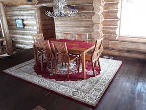 Uma mesa de jantar de madeira com seis cadeiras, cercada por um tapete estampado, em um interior de cabana rústica.