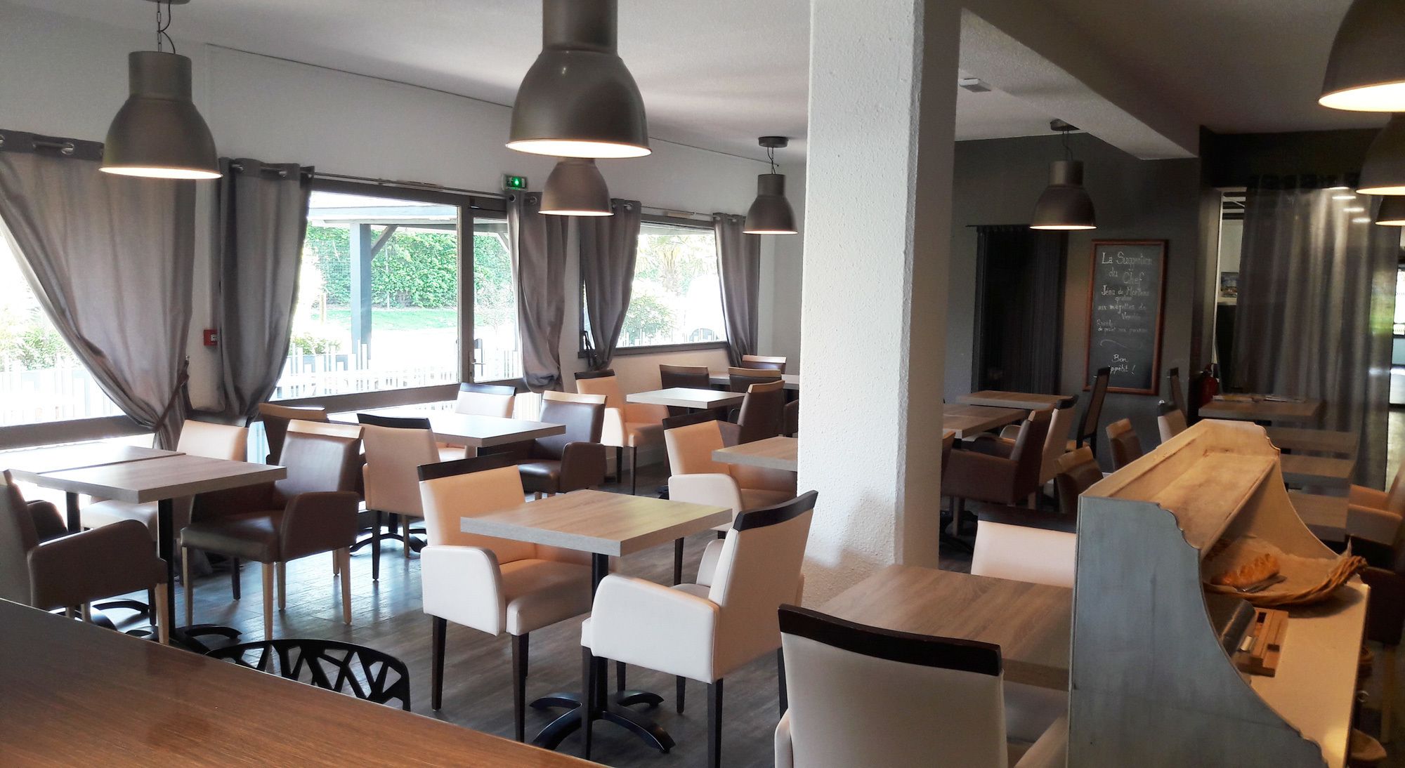 Un restaurant amb diverses taules i cadires, llums penjants grans i vistes a l'exterior per les finestres.