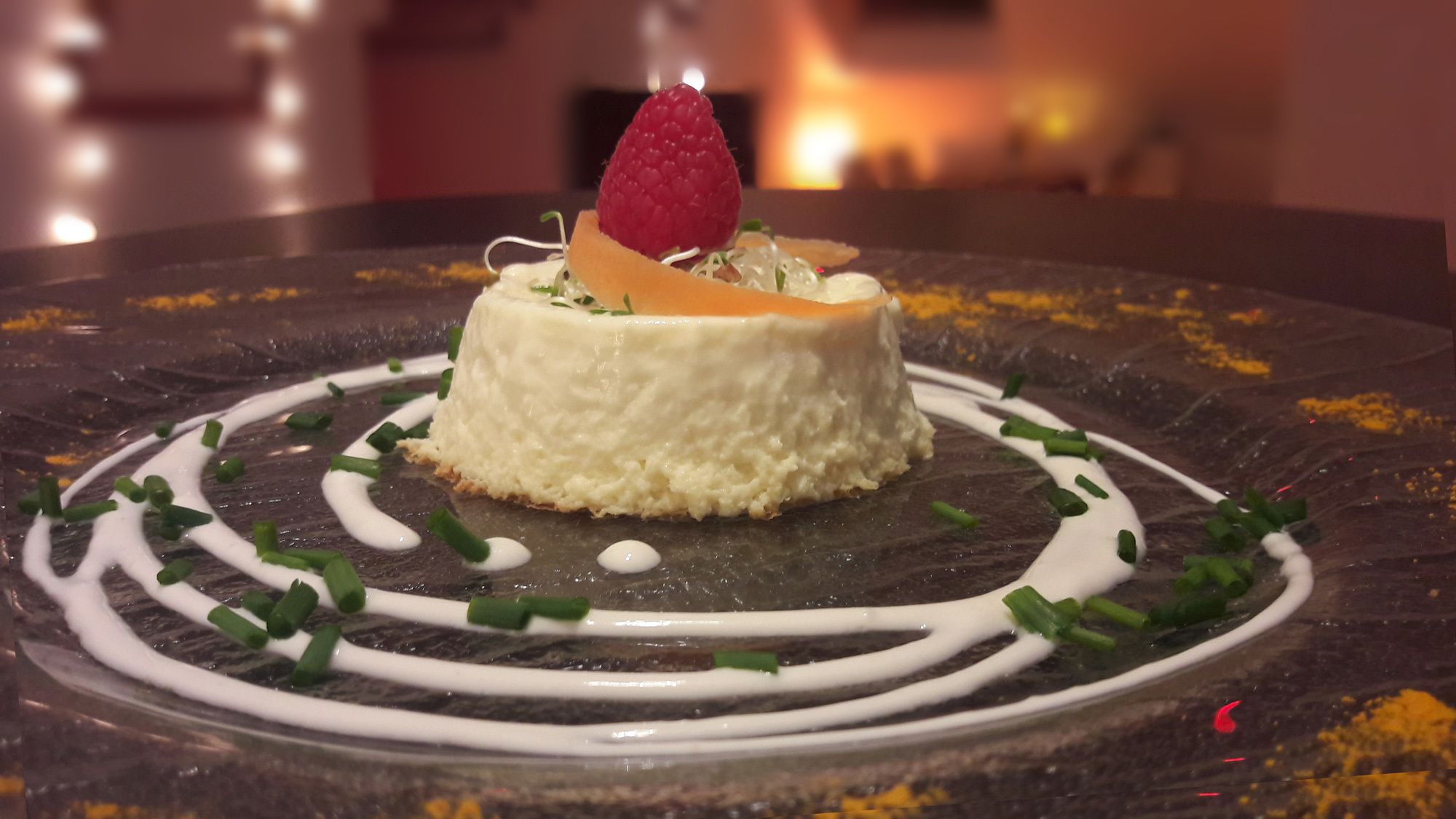 A dessert with a raspberry on top, surrounded by white sauce and green garnish on a dark plate.