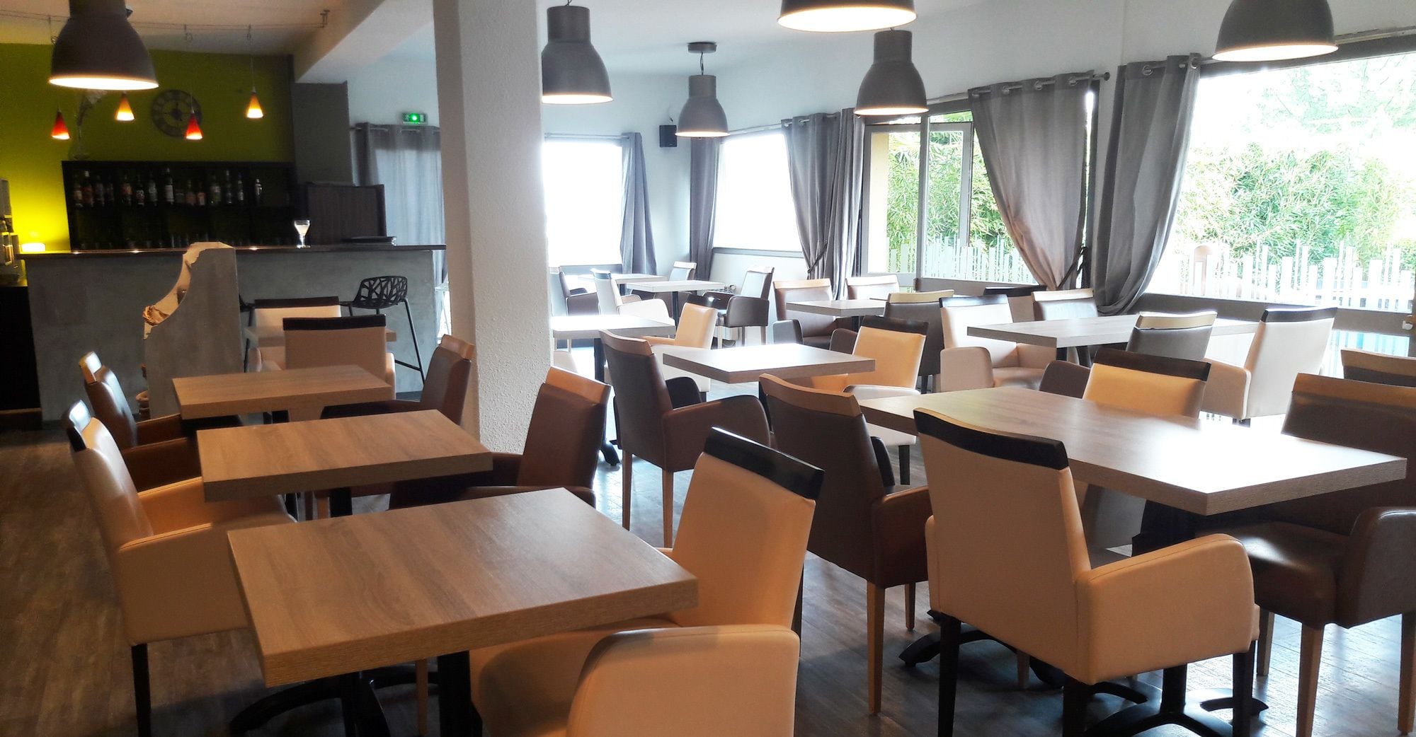 Un restaurante con varias mesas y sillas, una zona de bar y amplios ventanales con cortinas.
