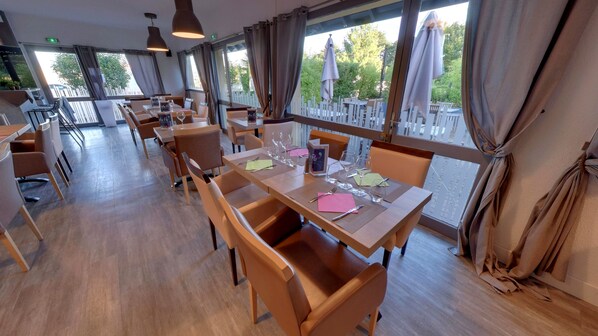 Un restaurante con mesas y sillas de madera, amplias ventanas con cortinas y sombrillas al aire libre.