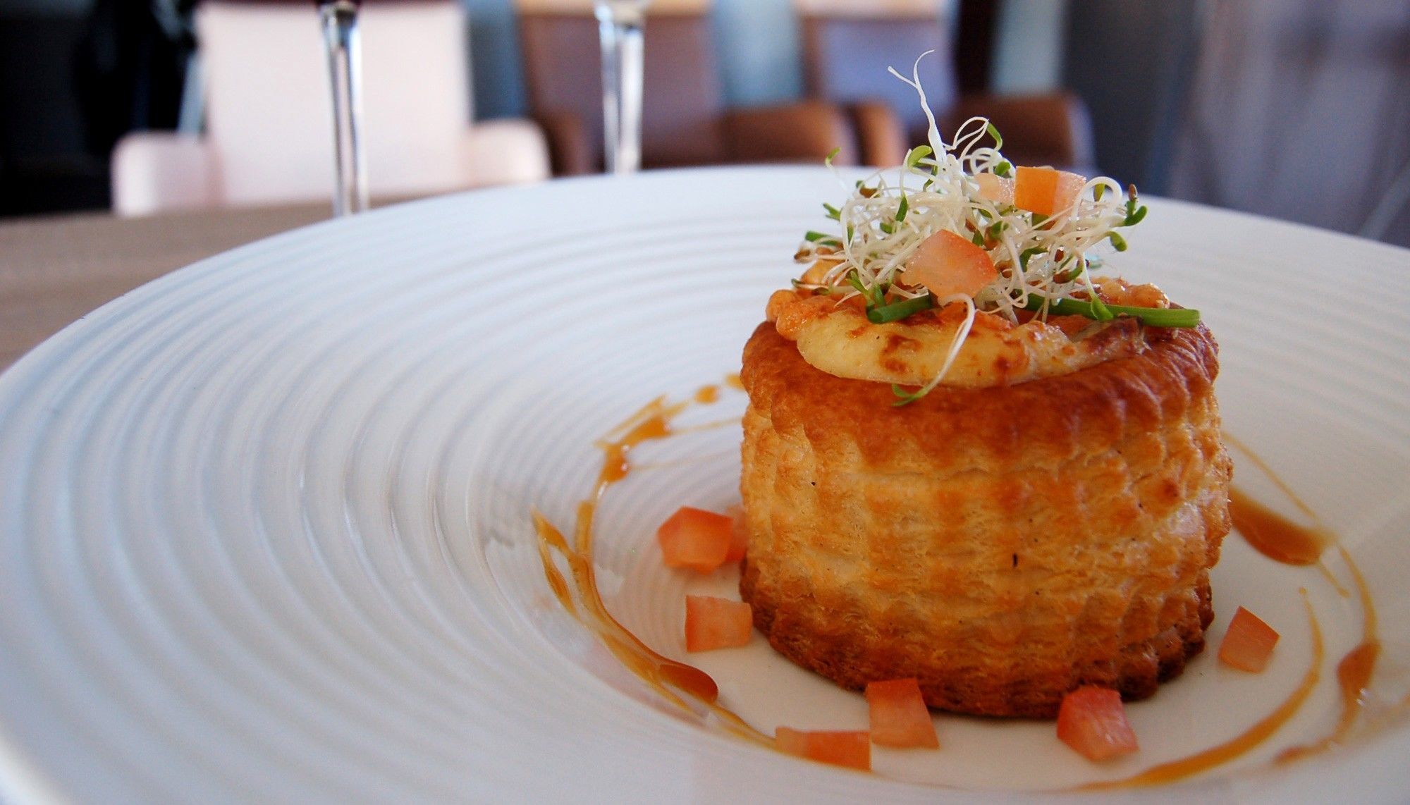 A plate with a food item, possibly a pastry, topped with sprouts and diced vegetables, served on a white plate with a textured rim.