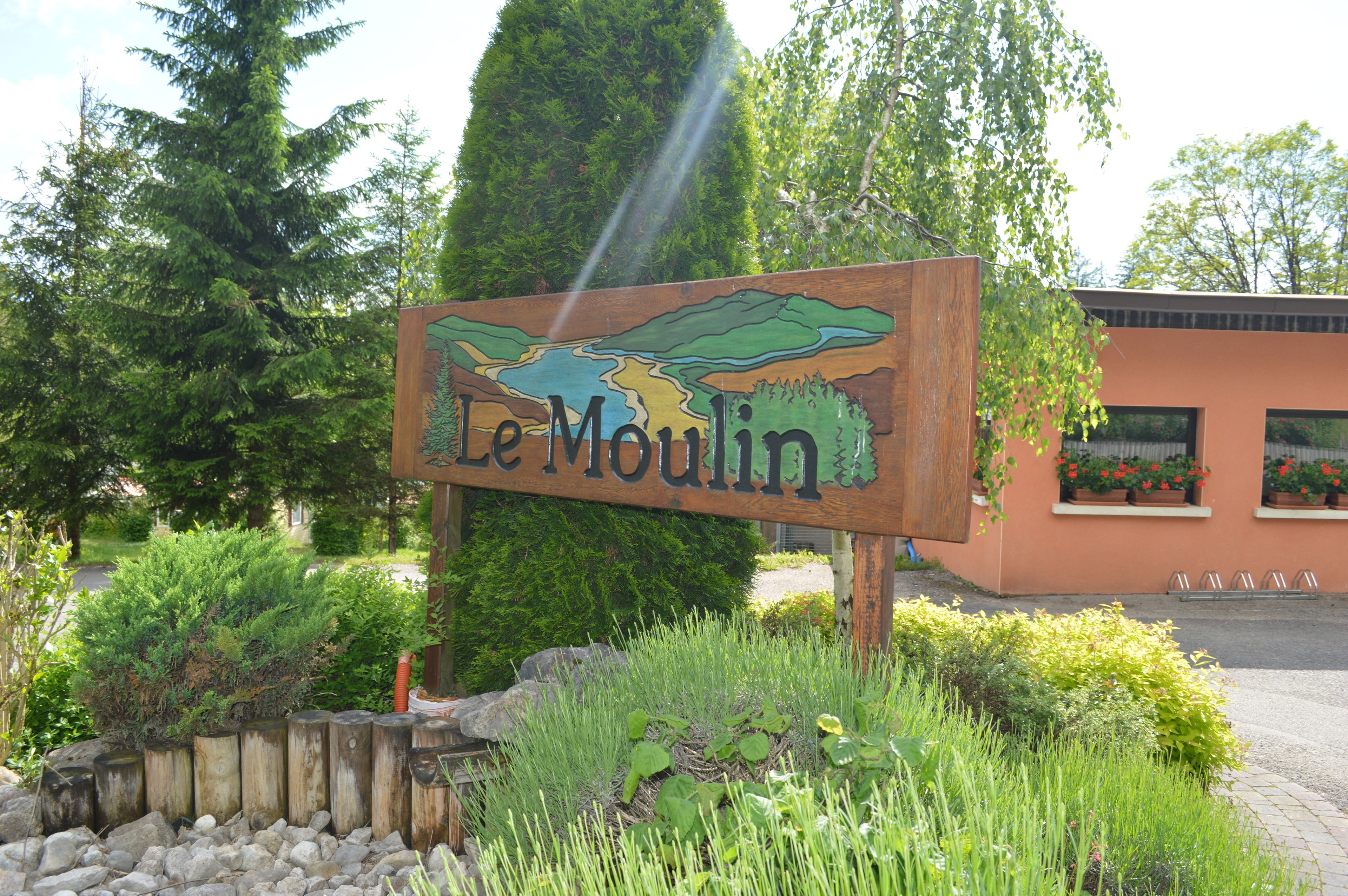 Ein hölzernes Schild mit der Aufschrift 'Le Moulin', umgeben von Grünpflanzen und einem Gebäude mit Fenstern.