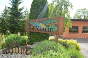 A wooden sign with 'Le Moulin' written on it, surrounded by greenery and a building with windows. - Camping Le Moulin (Patornay)