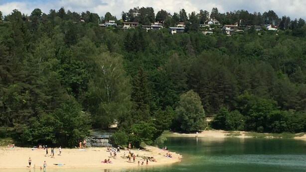 Udsigt over en sø med sandstrand, tæt skov og huse i baggrunden.