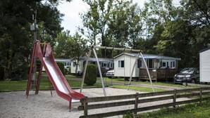 A playground with a red slide and swings, surrounded by a gravel area and a wooden fence. - Camping Les Pêcheurs (Pont-de-Poitte)