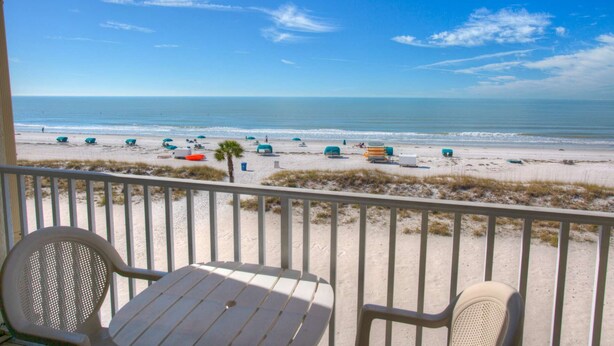 Blick auf einen Strand von einem Balkon mit Tisch und Stühlen, Strandsonnenschirmen und blauem Himmel.