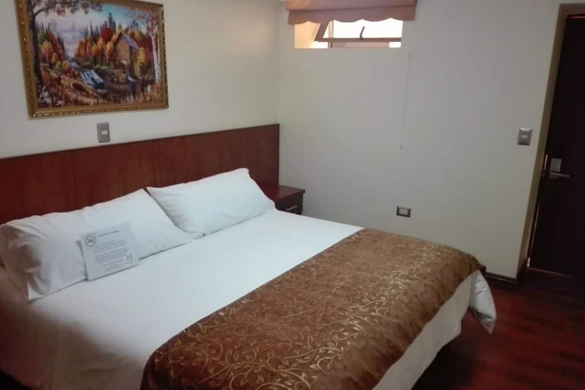 A hotel room with a large bed, a nightstand, a painting on the wall, and a window with curtains.