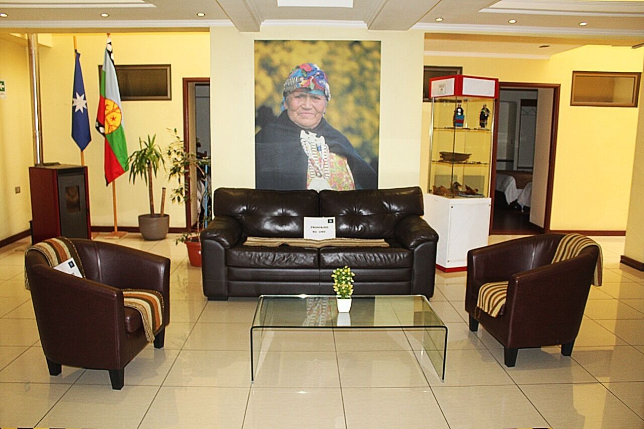 Un hall con un sofá de cuero, dos sillones, una mesa de centro de vidrio, un retrato y banderas.