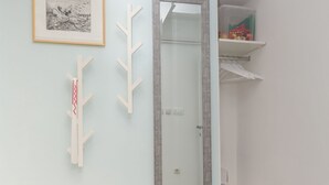 A room with a light blue wall, a framed black and white artwork, a wooden tree decoration, a mirror, and a white shelf with a red object. - Big Yard (Ljubljana)